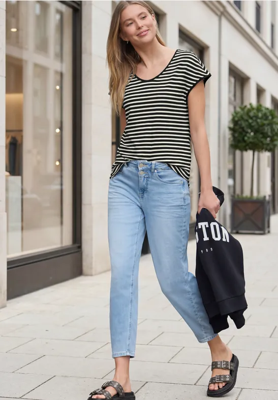 Frau in gestreiftem Shirt, Jeans und Sandalen schlendert am Gehweg und hält eine Jacke.