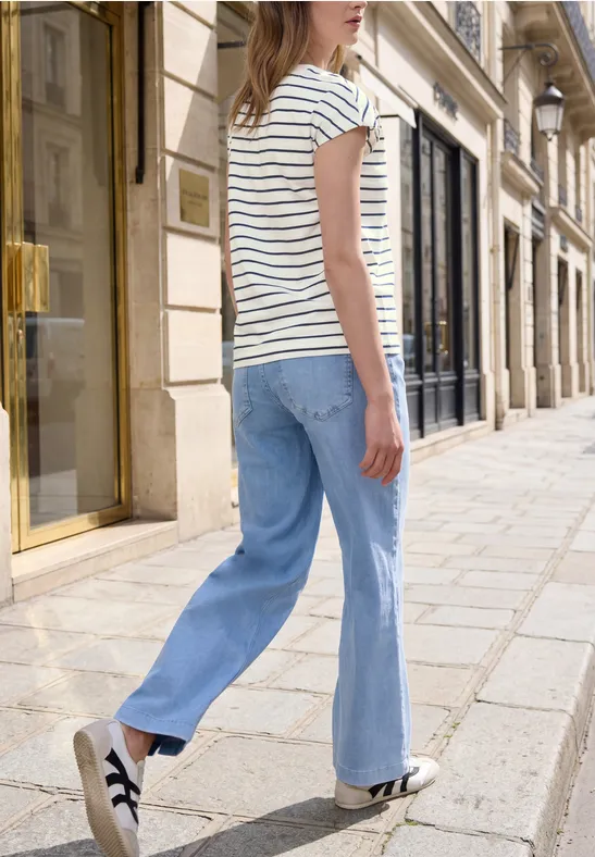 Frau in gestreiftem T-Shirt, blauer Jeans und Sneakern spaziert in einer schicken Fußgängerzone.