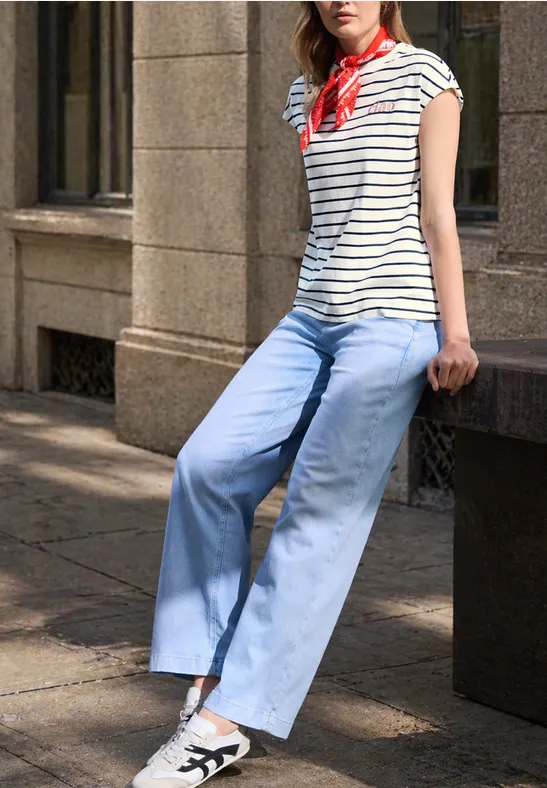 Person lehnt an einer Steinmauer, trägt weiß-blau gestreiftes Shirt, hellblaue weite Hose und rotes Halstuch.