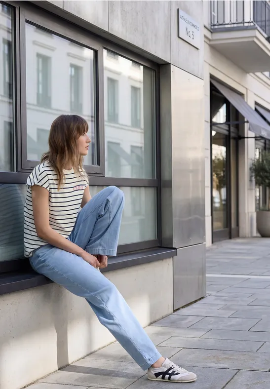 Eine junge Frau sitzt lässig auf einer Fensterbank an einer Hausfassade in der Stadt, trägt gestreiftes Shirt und Jeans.