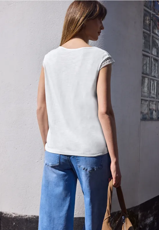 Vrouw van achteren in wit mouwloos topje en blauwe jeans, houdt een tas bij de muurhoek.