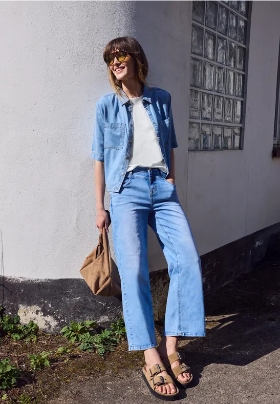 Een vrouw in jeans en sandalen leunt lachend tegen een muur, met zonnebril en schoudertas.