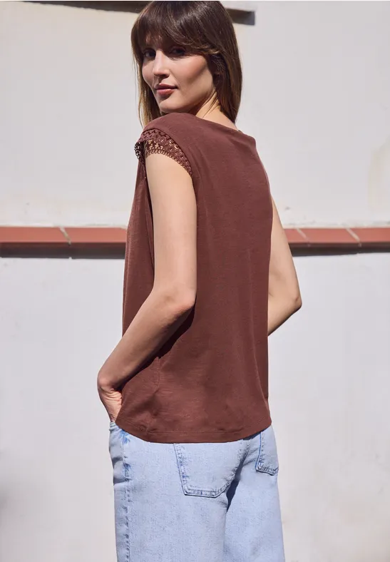 Vrouw in een mouwloos bruin topje en losse blauwe spijkerbroek, staand in profiel voor een witte muur.