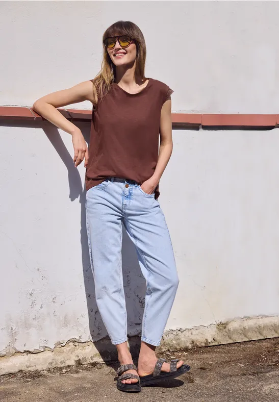 Persoon in bruin shirt, lichtblauwe jeans en sandalen leunt tegen witte muur met rode streep.