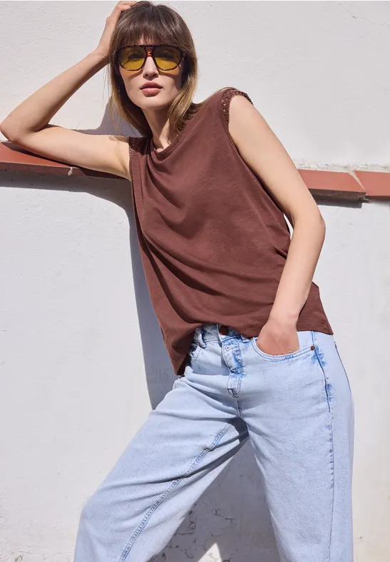 Vrouw leunt tegen een witte muur, draagt een bruin mouwloos topje en lichtblauwe jeans, hand in de zak.