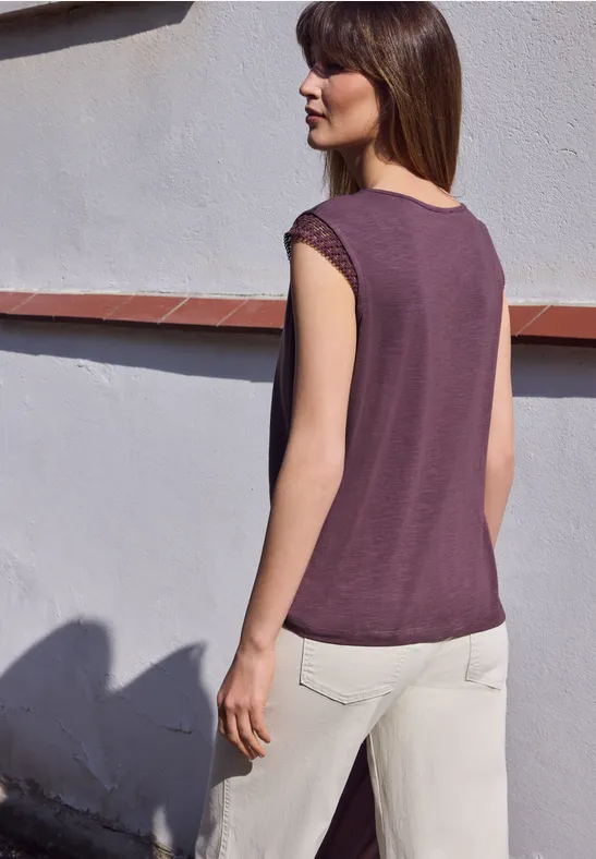 Frau in lila Top und hellen Hosen steht mit dem Rücken zu einer weißen Wand im Freien.