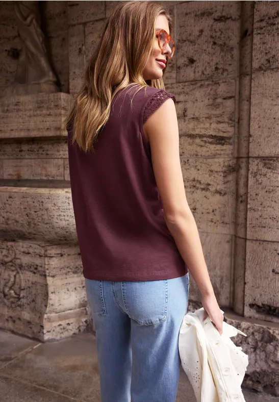 Vrouw van achter in een bordeauxkleurige mouwloze top en spijkerbroek, met een lichte jas voor een stenen muur.