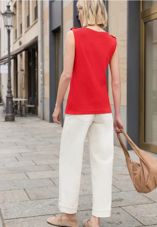 Vrouw in rood topje en witte broek met tas lopend op een geplaveide straat voor winkels.