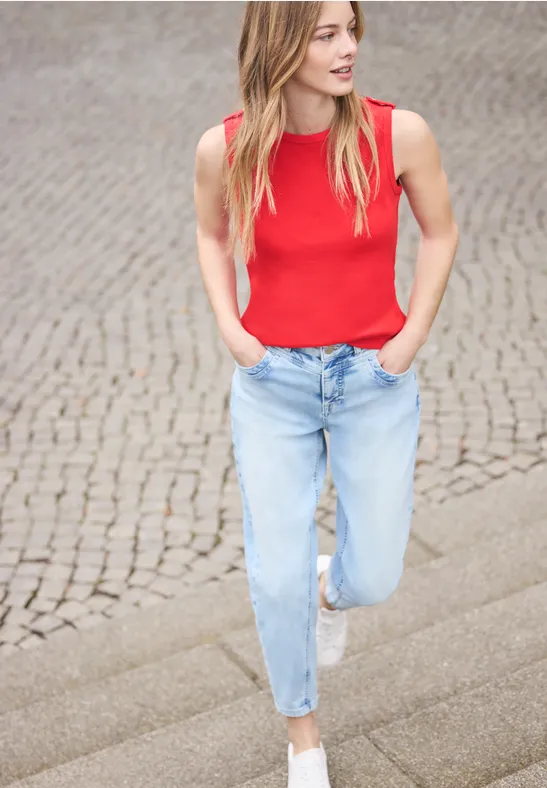 Vrouw in rood topje en lichtblauwe spijkerbroek loopt over een geplaveid pad.