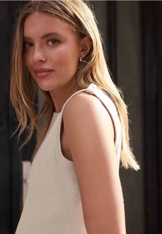 Młoda kobieta w profilu o blond włosach, ubrana w biały bezrękawnik na ciemnym tle.