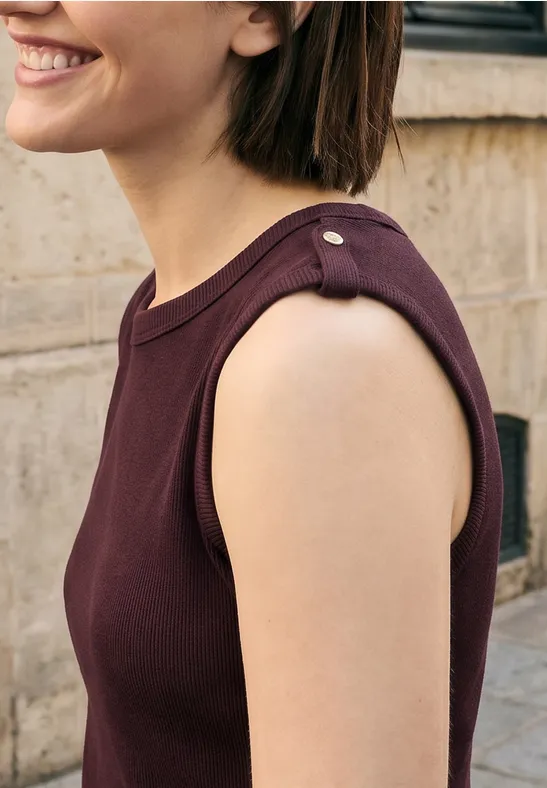 Portrait de profil d'une femme souriante en haut sans manches bordeaux devant un mur en pierre.