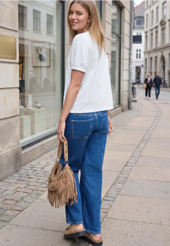 Frau mit weißem T-Shirt, weiten Jeans und brauner Fransen-Tasche schlendert vor Schaufenstern in der Innenstadt.