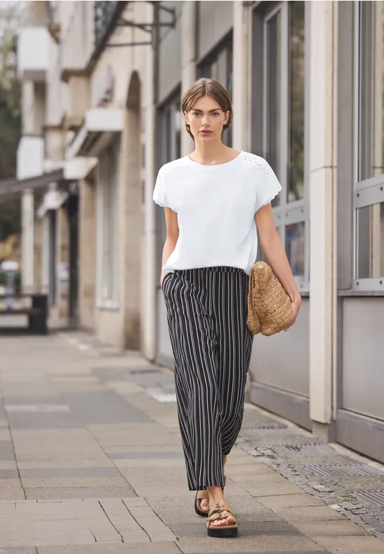 Vrouw in wit T-shirt en gestreepte broek wandelt door de stad met strooien tas en sandalen.