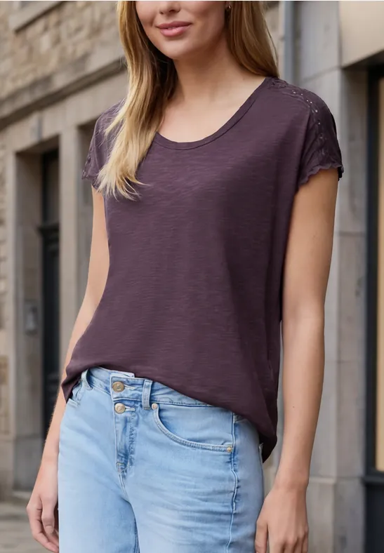 Frau trägt legeres lila T-Shirt und Jeans, steht vor einer Hauswand.