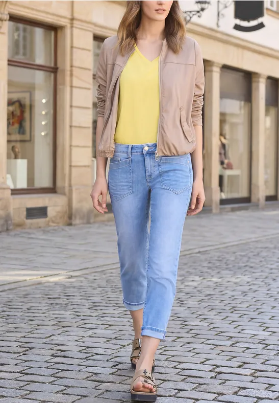 Frau geht in beiger Lederjacke, gelbem Top, hellen Jeans und Sandalen über Kopfsteinpflaster.