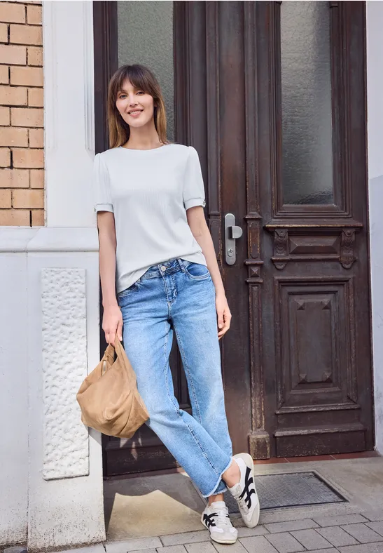 Eine Frau mit weißem T-Shirt und Jeans lehnt lässig an einer Wand vor einer Holztür und hält eine Tasche.