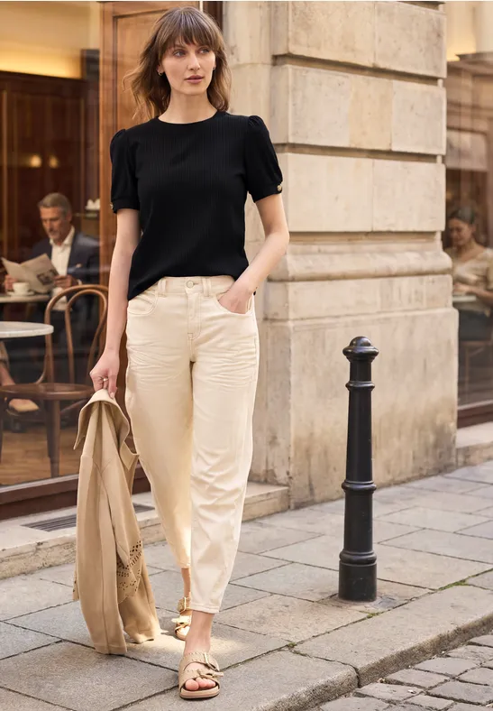 Eine Frau in schwarzem T-Shirt und beigen Hosen mit Jacke in der Hand spaziert an einem Café vorbei.