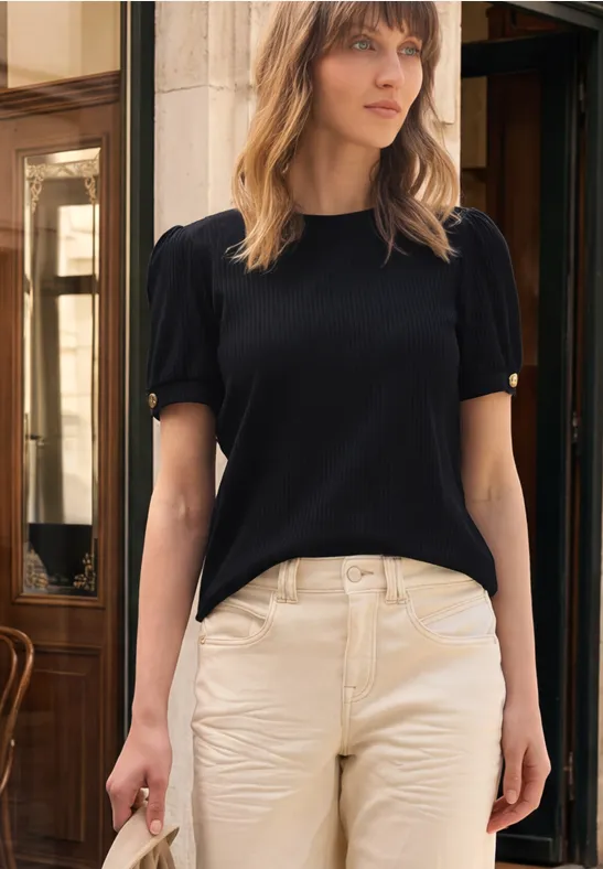 Frau trägt schwarzen Kurzarmpullover und helle Hose, steht vor einer Holztür in Café-Atmosphäre.