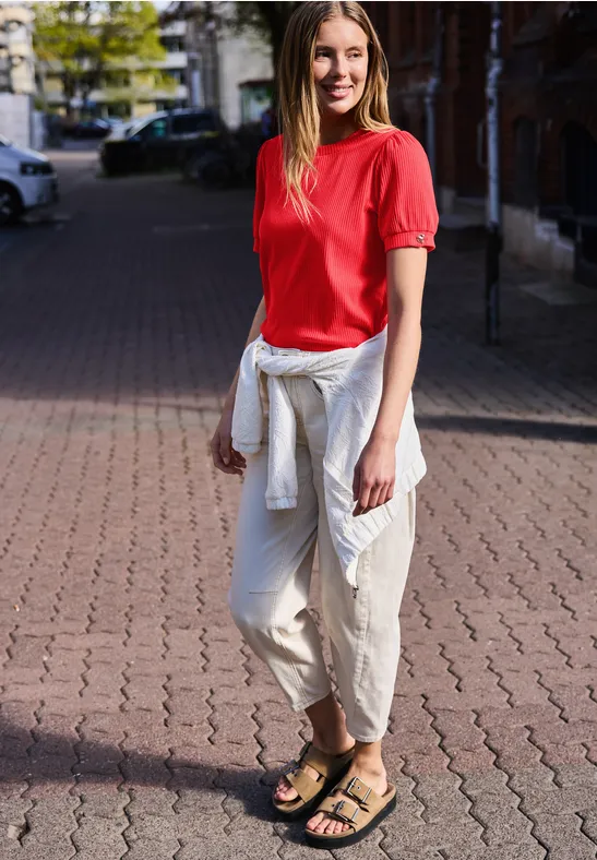 Person steht auf gepflastertem Gehweg, trägt rotes Oberteil, weiße Hose, weiße Jacke um die Hüfte und Sandalen.