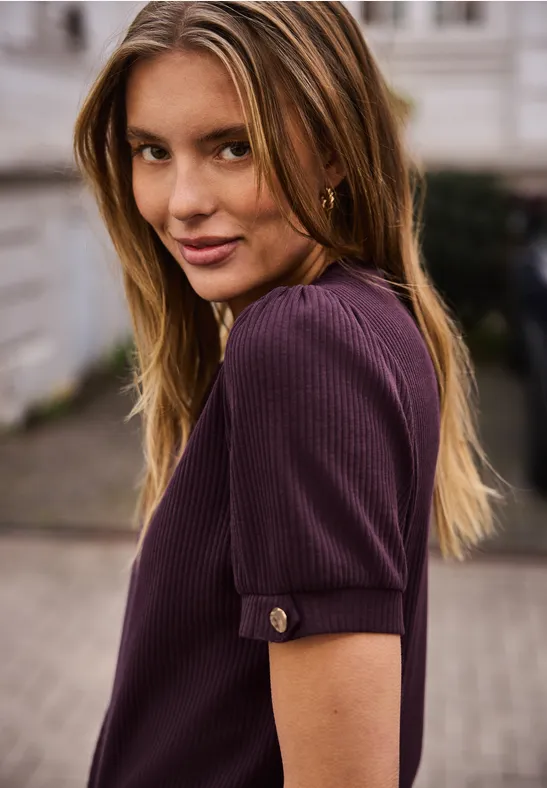 Jonge vrouw met lang haar in paarse korte mouw top, kijkt over haar schouder tegen stedelijke achtergrond.