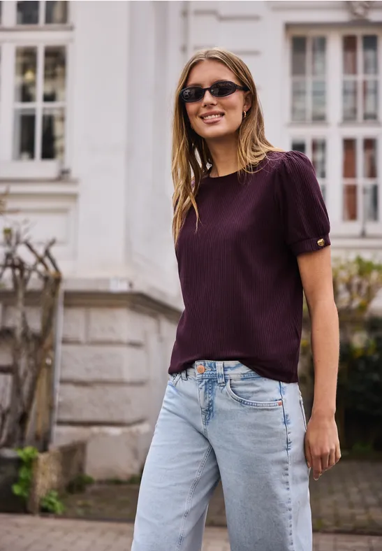 Vrouw in een paarse blouse en lichte jeans staat voor een huis met witte ramen.