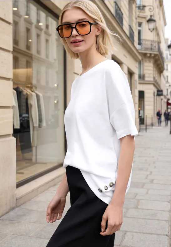 Frau in weißem T-Shirt und schwarzer Hose mit Knöpfen läuft über eine gepflasterte Einkaufsstraße.