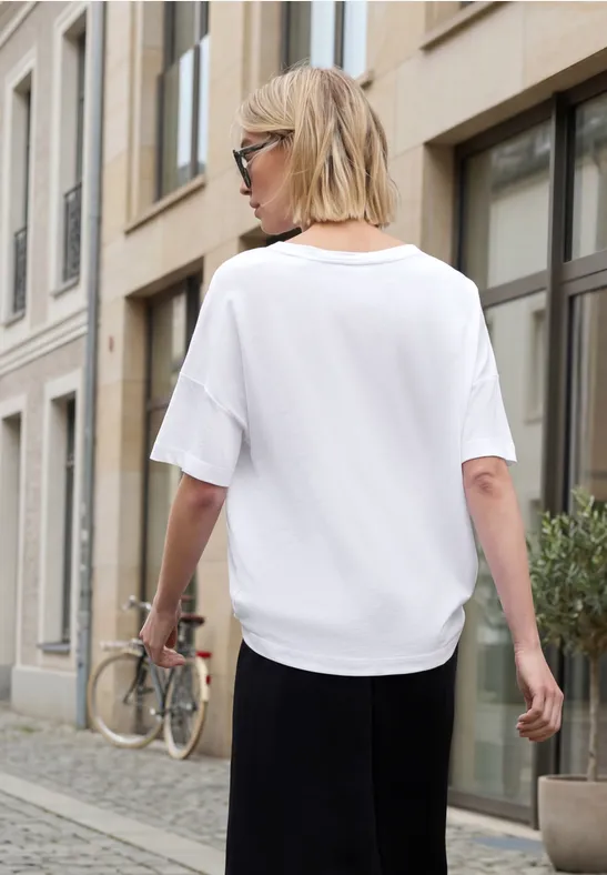 Eine Frau in weißem T-Shirt, schwarzer Rock und Sonnenbrille geht an einem Stadthaus mit Fahrrad vorbei.