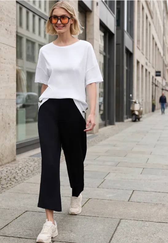 Eine Frau im weißen T-Shirt, schwarzen Hosen und weißen Sneakers läuft in der Stadt auf dem Gehweg.