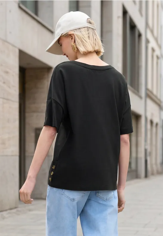 Frau mit schwarzem T-Shirt, Jeans und weißer Cap, von hinten auf städtischem Gehweg vor modernem Gebäude.