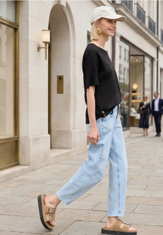 Frau in schwarzem T-Shirt, hellen Jeans und Sandalen schlendert an schicken Geschäften vorbei.