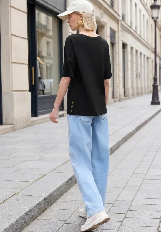 Eine Frau in schwarzem T-Shirt, heller Jeans und weißer Kappe geht auf Kopfsteinpflaster an Stadthäusern vorbei.