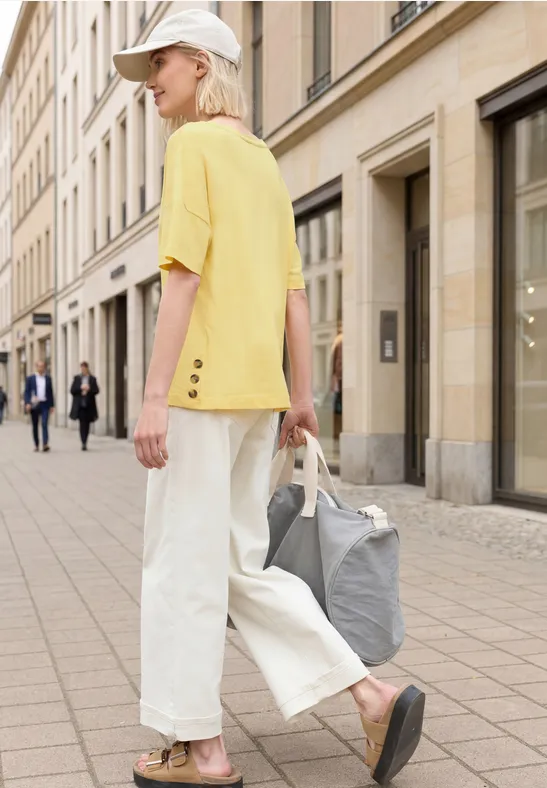 Eine Person mit gelbem T-Shirt, weißen weiten Hosen, Cap und grauer Tasche läuft über den Bürgersteig.