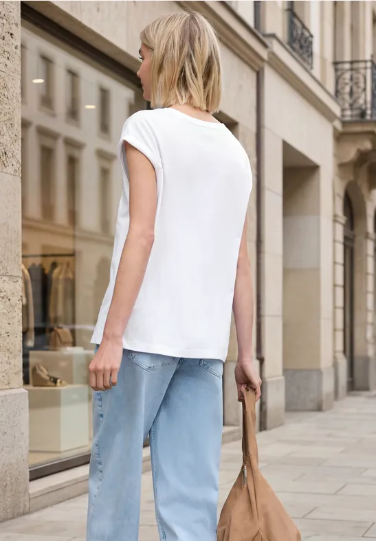 Vrouw met wit T-shirt, lichte jeans en tas wandelt door een chique stadsstraat.