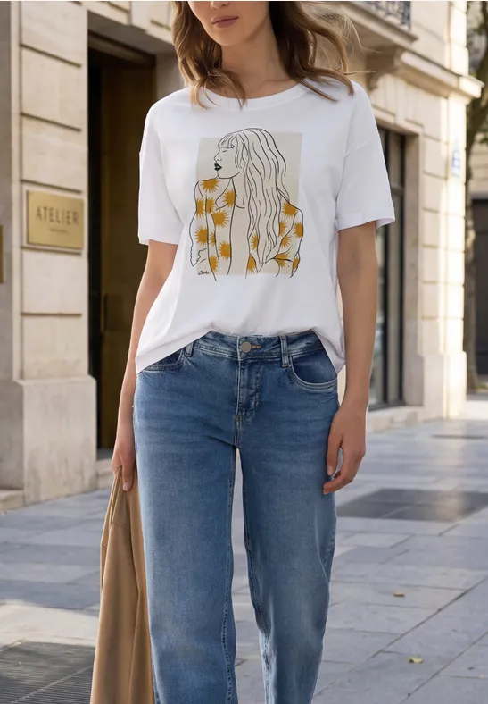 Frau trägt weißes T-Shirt mit abstraktem Frauenporträt und gelbem Muster, kombiniert mit blauen Jeans auf einer Stadtstraße.