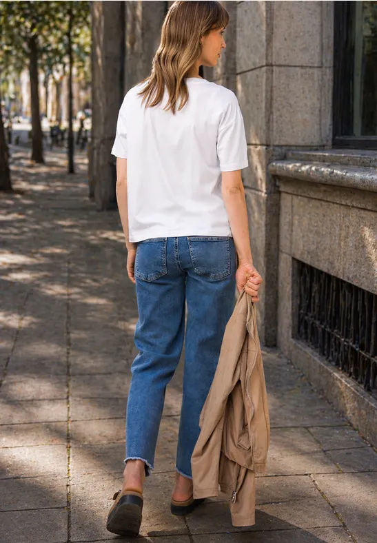 Frau in weißem T-Shirt und blauen Jeans geht mit hellbrauner Jacke in der Hand eine sonnige Straße entlang.