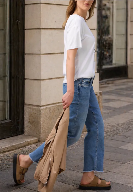 Frau in weißem T-Shirt, blauen Jeans und braunen Sandalen trägt eine beige Jacke auf dem Gehweg.