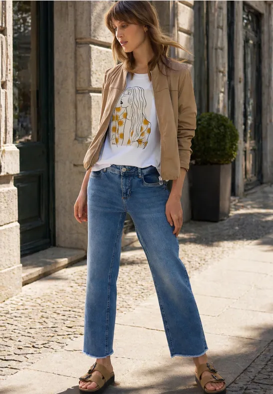 Frau in beiger Jacke, weißem Print-T-Shirt und weiten, knöchellangen Jeans steht auf einem Bürgersteig vor einem Gebäude.