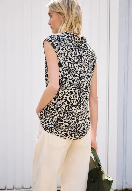Eine Frau von hinten in floral gemustertem Top, hellen Hosen und grünem Beutel vor heller Wand.
