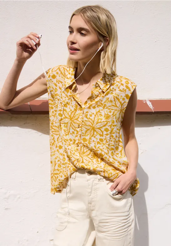 Vrouw voor lichte muur in gele bloemenblouse en witte broek.
