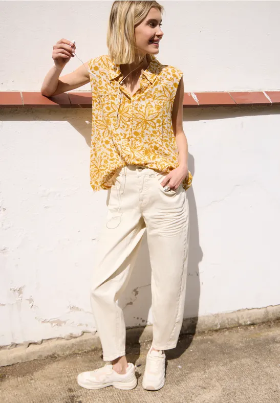 Persoon leunt tegen een witte muur, draagt een gele top met print, lichte broek en witte sneakers.