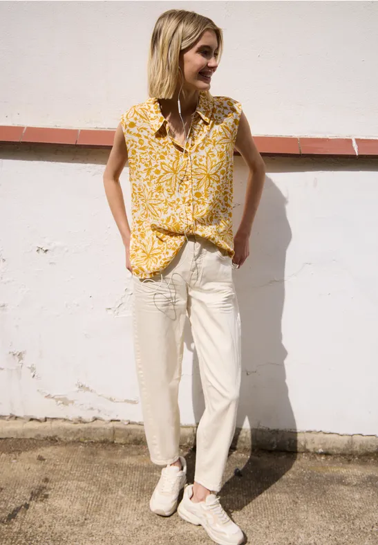 Persoon leunt tegen lichte muur, draagt gele gebloesde top, lichte broek en witte sneakers.