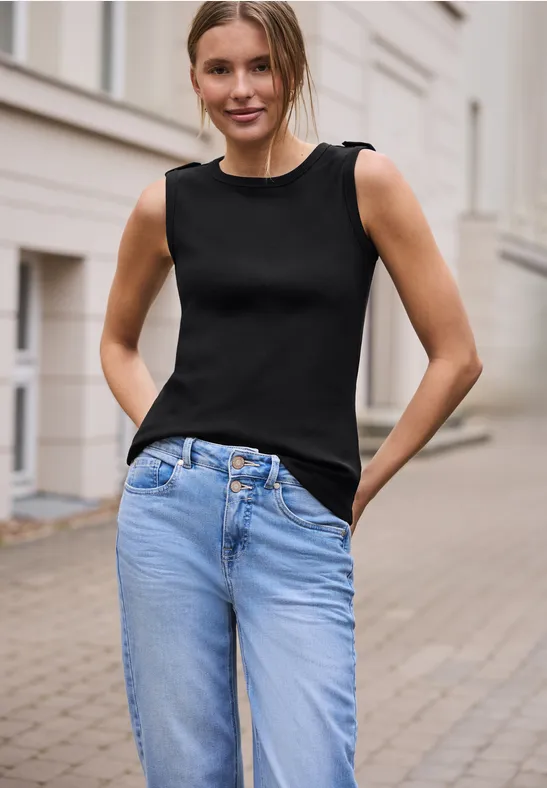 Vrouw in zwarte mouwloze top en blauwe jeans staat ontspannen op een geplaveide straat voor een licht gebouw