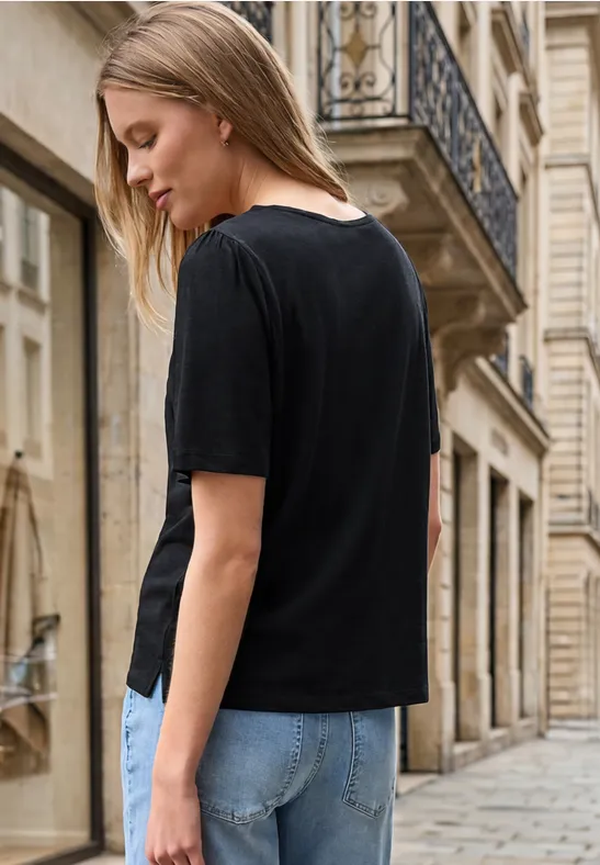 Eine Frau in schwarzem T-Shirt und blauen Jeans steht seitlich auf einer gepflasterten Straße vor historischen Gebäuden.