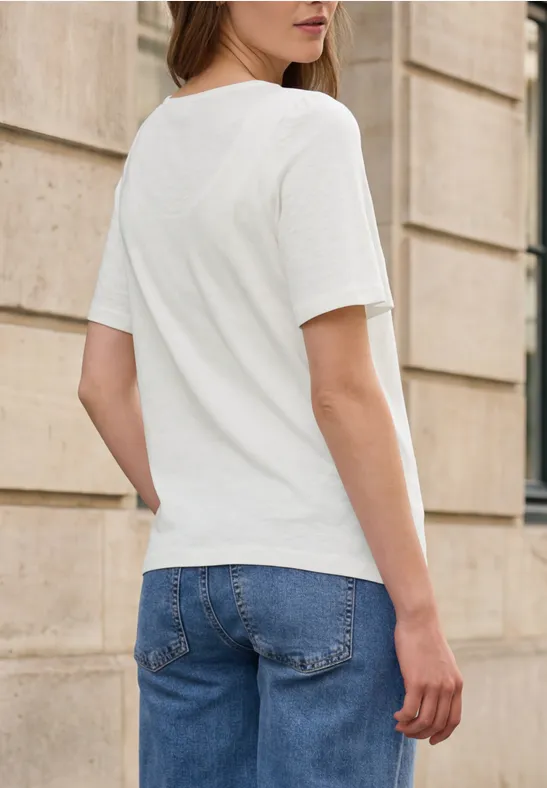 Frau in weißem T-Shirt und blauen Jeans, steht mit dem Rücken zur Kamera vor einer beigen Steinwand.