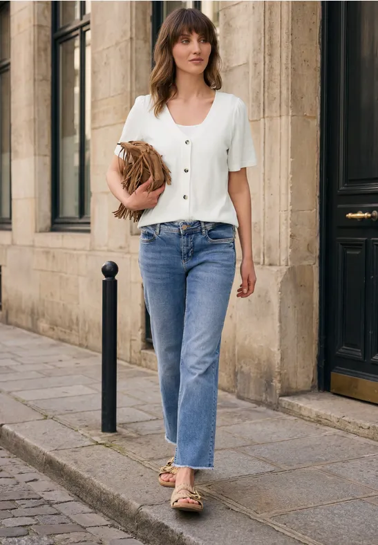 Person mit Jeans, weißem Shirt, Sandalen und Strohtasche geht an einer Hausfassade entlang.