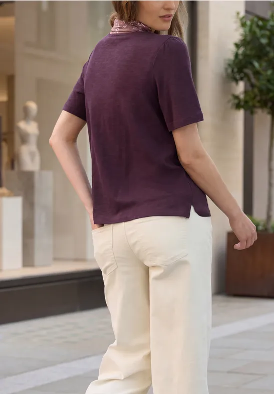 Frau mit lila Kurzarmshirt und hellen Hosen schlendert in einer städtischen Einkaufsstraße.