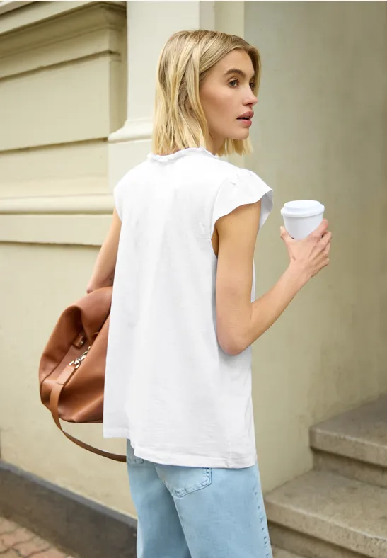 Persoon van achteren met wit T-shirt, jeans en koffiebeker, met een bruine tas voor een gevel.