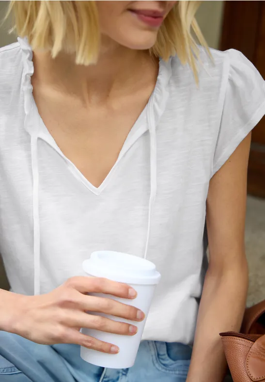Vrouw in wit shirt houdt koffiebeker vast, zittend met tas naast zich.