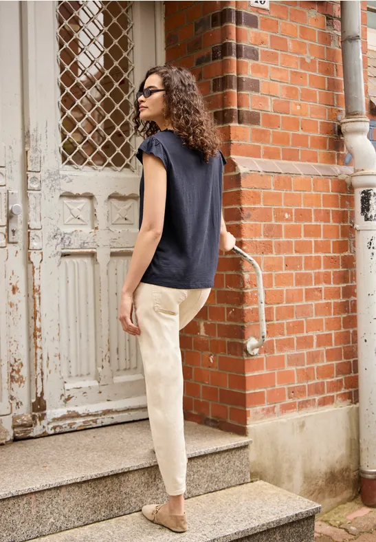 Vrouw met krullend haar en zonnebril, donkerblauw shirt, lichte broek en beige tas beklimt een trap.