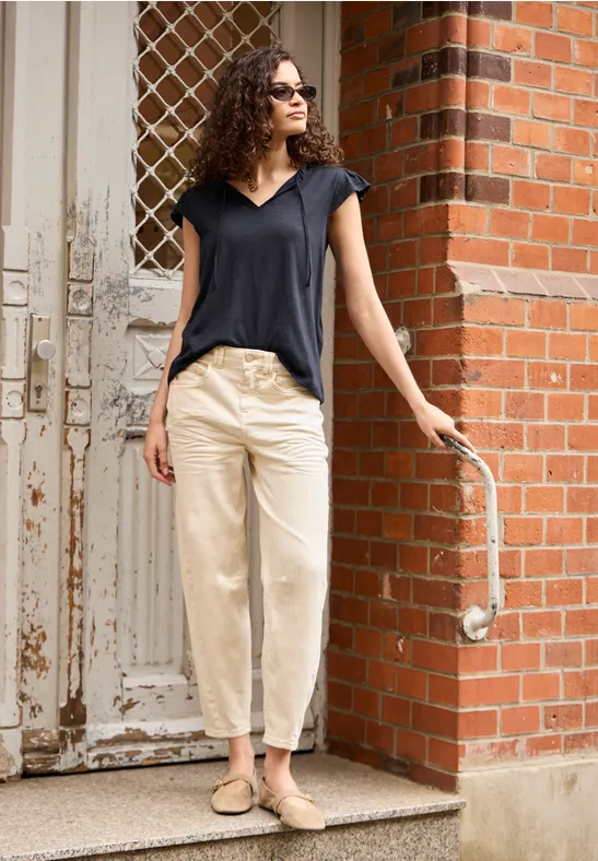 Vrouw leunt nonchalant tegen bakstenen muur, draagt zwart shirt, lichte broek, instappers en zonnebril.